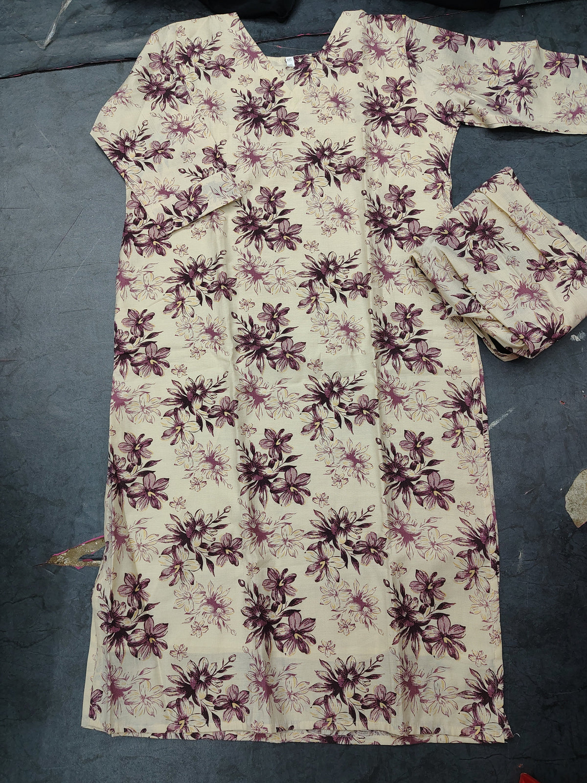 A cream-colored long kurta with a dark purple floral pattern, laid flat on a dark gray floor next to matching pleated fabric.