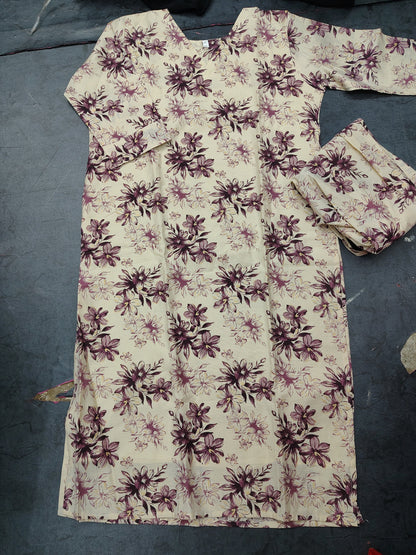 A cream-colored long kurta with a dark purple floral pattern, laid flat on a dark gray floor next to matching pleated fabric.